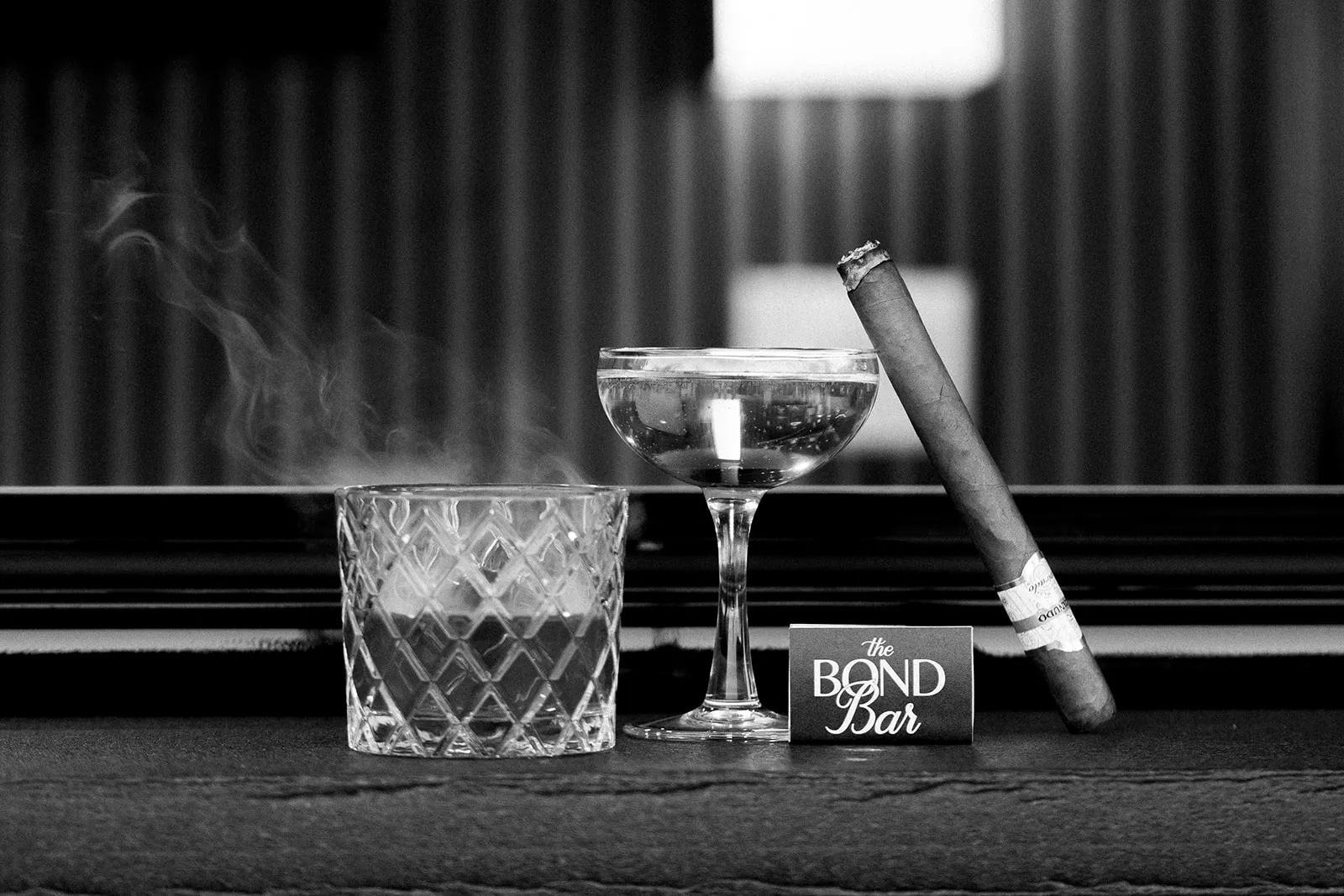 Black and white still life of a crafted cocktail, cigar, and glassware representing luxury mobile cigar and cocktail entertaining