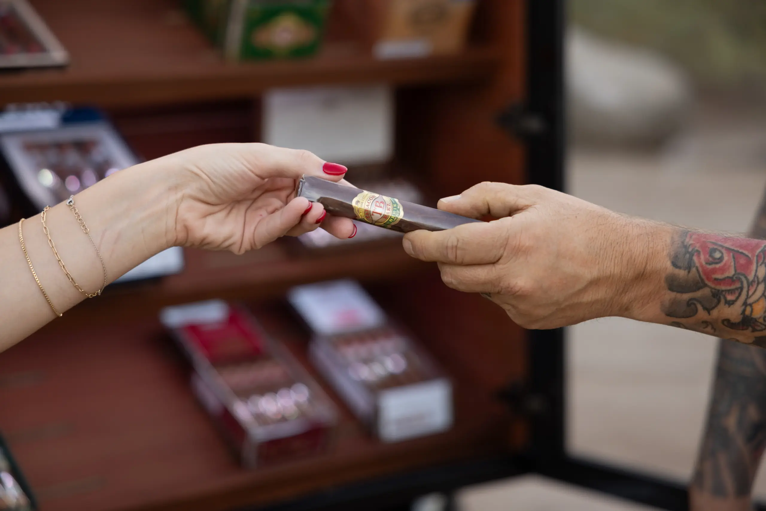 Two hands exchange a small, cigar-shaped wrapped item between a person with red nails and a tattooed forearm.
