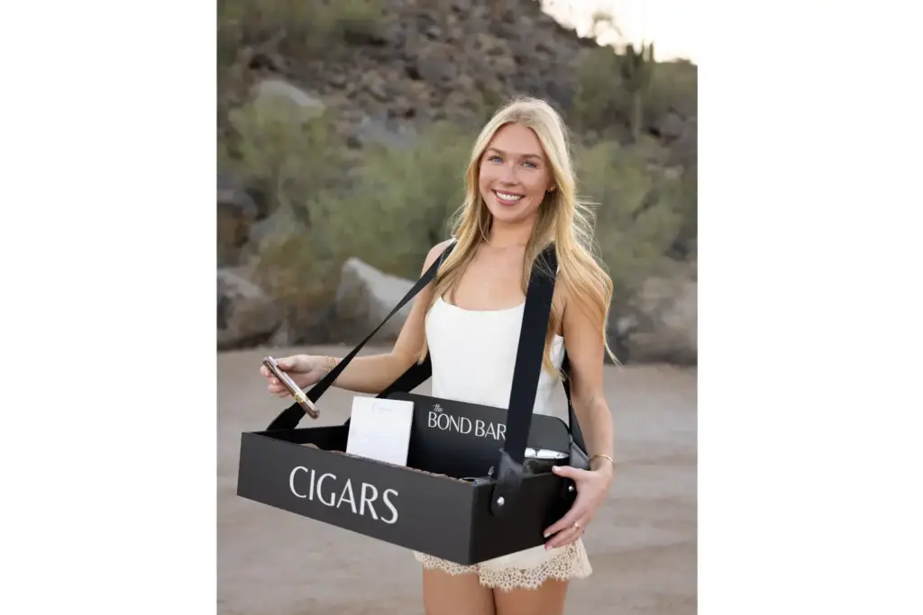 Young woman outdoors holding a black box labeled CIGARS, smiling at the camera.