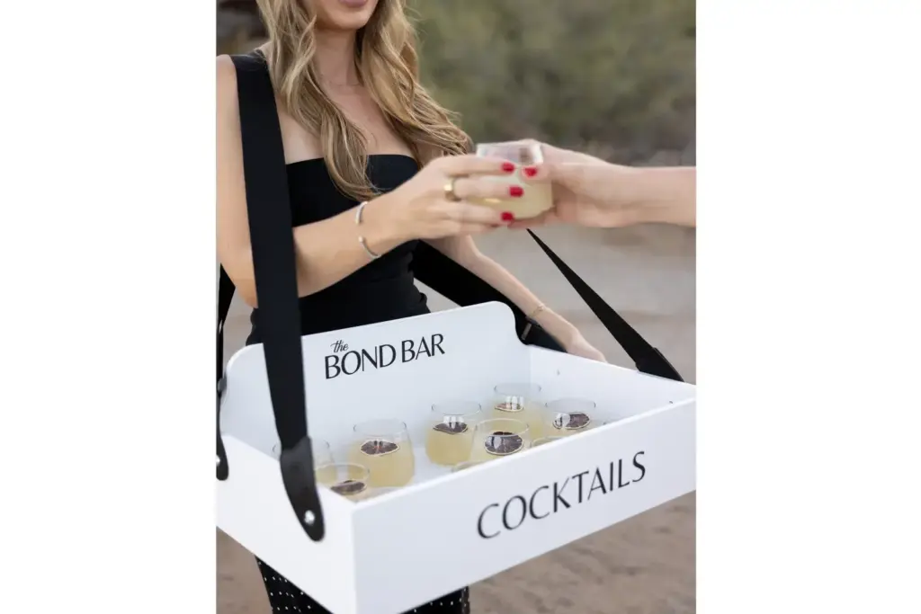 Woman in a black dress holds a white cocktail box labeled "COCKTAILS" with small glasses inside, handing a drink to someone off-frame.
