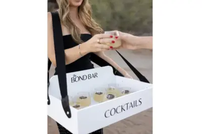 Woman in a black dress holds a white cocktail box labeled "COCKTAILS" with small glasses inside, handing a drink to someone off-frame.