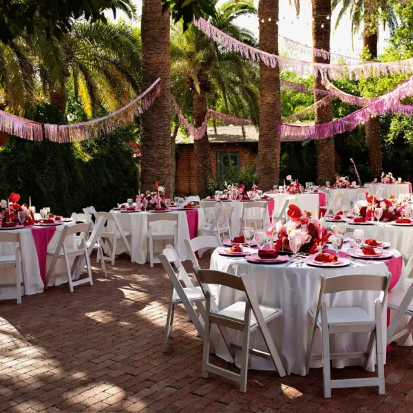 Outdoor garden wedding reception with pink decor, round tables, white chairs, and palm trees with string lights in the background.