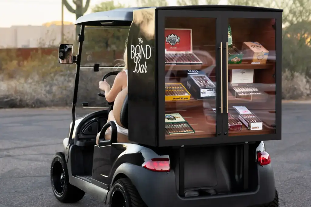 Golf cart with a black, cigar humidor cabinet labeled 'The Bond Bar' mounted on the back, displaying cigars through glass doors